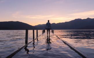 Silhouette of a person from behind standing at the end of a dock looking out at a lake. The sun is rising behind the mountains.